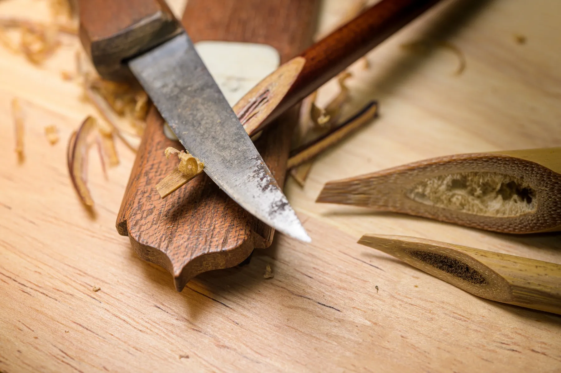 A knife carving the nib of a reed calligraphy pen on a wooden cutting block, with shavings scattered and two split reed nibs showing their hollow interiors alongside.